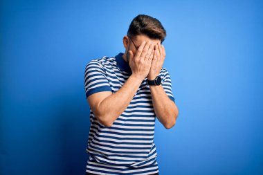 Mavi gözlü, gözlüklü, mavi arkaplanlı, mavi çizgili tişörtlü, suratını ellerle örten ve ağlayan genç bir adam. Depresyon kavramı.