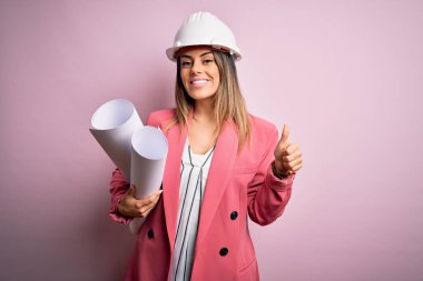 Genç, güzel, esmer, mimar, güvenlik kaskı takan, elinde planlar olan mutlu, gülümseyen, işaret veren, parmaklarıyla başparmağını kaldıran bir kadın.