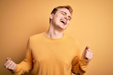 Sarı arka plan üzerine günlük süveter giyen genç ve yakışıklı bir adam mutlu ve heyecanlı bir şekilde kollarını kaldırmış, gülümseyen ve başarı çığlıkları atan bir jest yapıyor. Kutlama konsepti.