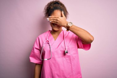 Afro-Amerikalı hemşire kız tıbbi üniforma ve steteskop takmış pembe arka planda gülümsüyor ve yüzünde şaşkınlıktan gözleri kapayarak gülüyor. Kör kavram.