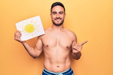 Tatilde sakallı, mayo giyen, elinde güneş kâğıdıyla gülümseyen, mutlu, eli ve parmağı yanda olan yakışıklı bir adam.