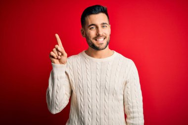 İzole edilmiş kırmızı arka plan üzerinde duran, rahat beyaz süveter giyen genç yakışıklı bir numaralı parmağını gösterirken kendinden emin ve mutlu bir şekilde gülümsüyor..