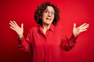Orta yaşlı, güzel, kıvırcık saçlı bir kadın kırmızı arka planda gözlük takıyor. Kolları ve elleri havada, kafası karışmış bir ifade takınıyor. Şüphe kavramı.
