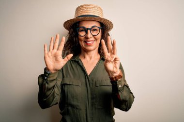 Orta yaşlı esmer kadın gözlüklü ve şapkalı. İzole edilmiş beyaz arka planda duruyor. Sekiz numaralı parmaklarıyla işaret ediyor..
