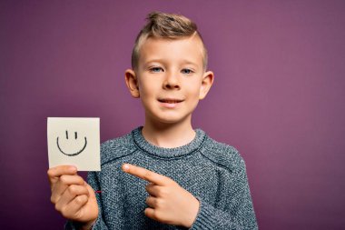 Küçük beyaz bir çocuk kağıtta gülen yüzünü gösteriyor. Mutlu bir mesaj. El ve parmak ile göstererek çok mutlu.