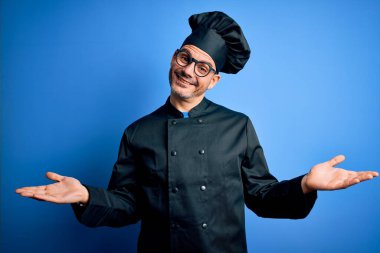Genç, yakışıklı aşçı üniformalı ve şapkalı, mavi arka planda gülümseyen, her iki elini de açık gösteren, sunan ve reklam karşılaştırma ve denge