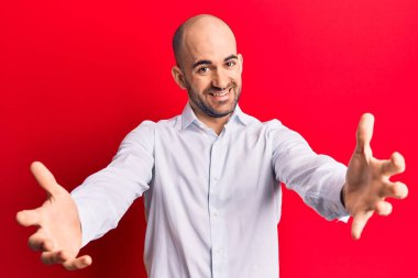 Zarif bir gömlek giymiş genç ve kel bir adam kameraya bakıp sarılmak için kollarını açmış gülümsüyor. Mutluluğu kucaklayan neşeli bir ifade.. 