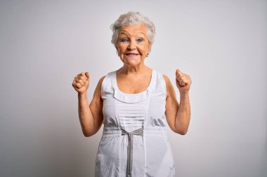 Son sınıf güzeli gri saçlı kadın beyaz arka plan üzerine günlük yazlık elbise giyip kollarını kaldırarak ve açık gözlerle başarının şaşkınlığını ve şaşkınlığını yaşıyor. Kazanan konsept.