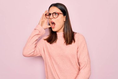 Genç, güzel, esmer bir kadın pembe arka planda günlük süveter ve gözlük takıp el ele tutuşarak bağırıyor. İletişim konsepti.