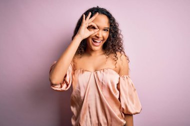 Kıvırcık saçlı, pembe tişörtlü genç bir kadın pembe arka planda dikiliyor. El hareketleriyle gülümsüyor, gözleriyle mutlu yüzüyle parmaklarının arasından bakıyor..