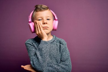 Kulaklık takan küçük beyaz bir çocuk mor arka planda müzik dinliyor. Kollarını kavuşturup depresyondan sıkılmış ve yorgun görünüyor..