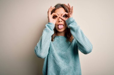 İzole edilmiş arka planda günlük mavi süveter giyen genç sarışın kız dürbün gibi dil çıkartıyor, gözler parmaklarından bakıyor. Çılgın ifade.