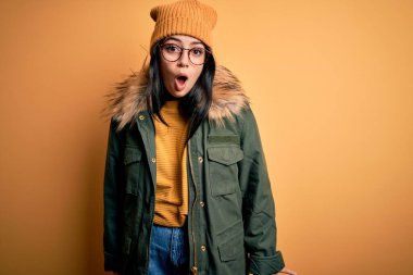 Genç esmer kadın, gözlük takıyor ve sarı şapkalı kışlık ceket giyiyor. Arka planda, korku, korku ve heyecan dolu bir ifade var..