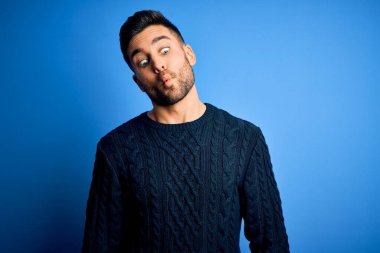 İzole edilmiş mavi arka plan üzerinde duran sıradan süveter giyen genç yakışıklı adam dudaklı balık suratı yapıyor, çılgınca ve komik bir jest. Komik ifade.