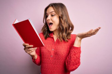 Pembe izole edilmiş arka plan üzerine kitap okuyan genç sarışın kız çok mutlu ve heyecanlı, kazanan yüz ifadesi zaferi büyük bir gülümsemeyle kutluyor ve ellerini kaldırıyor.