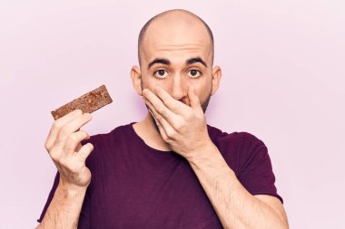 Genç, kel, enerji proteini barını tutan, ağzı elle kaplayan, şok olmuş ve hata yapmaktan korkan genç bir adam. şaşırmış ifade 