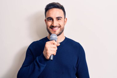 Beyaz arka planda mikrofon kullanarak şarkı söyleyen sakallı yakışıklı genç şarkıcı pozitif ve mutlu görünüyor ve kendine güvenen bir gülümsemeyle dişlerini gösteriyor.