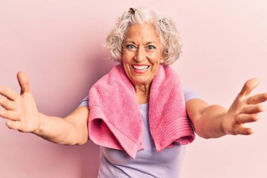 Spor kıyafetleri ve havlu giymiş yaşlı gri saçlı kadın kucaklaşmak için kollarını açmış kameraya bakıyor. Mutluluğu kucaklayan neşeli bir ifade.. 