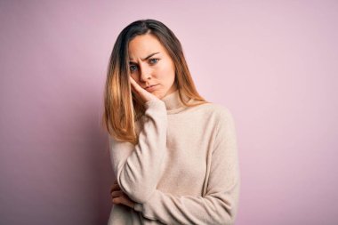 Mavi gözlü güzel sarışın kadın pembe arka planda balıkçı yaka kazak giyiyor ve kollarını kavuşturarak depresyondan sıkılmış ve yorgun görünüyor..
