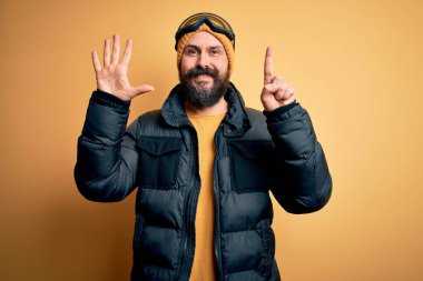 Sakallı, kar maskesi takmış, kar maskesi takmış, kel yakışıklı kayakçı altı numaralı parmaklarıyla kendini beğenmiş ve mutlu gülümseyen..