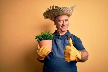 Orta yaşlı, beyaz önlüklü ve şapkalı bir çiftçi sarı arka planda saksı tutuyor, gülümsüyor ve gülümsüyor.