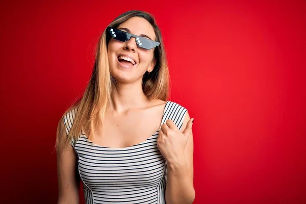 Young beautiful brunette woman wearing funny thug life sunglasses over red background smiling with happy face looking and pointing to the side with thumb up.