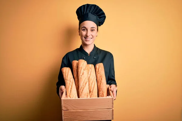 炊飯器の制服を着た若い美しいパン屋の女性と幸せな顔をしたパンで木の箱を保持し 歯を示す自信を持って笑顔で笑みを浮かべて — ストック写真