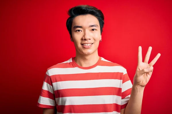 若いですハンサムな中国人男性身に着けていますカジュアルストライプTシャツ上に立って赤い背景を示すと指の数3で指摘しながら 自信と幸せ — ストック写真