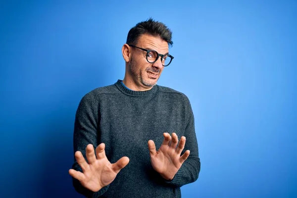 Young handsome man wearing casual sweater and glasses standing over blue background disgusted expression, displeased and fearful doing disgust face because aversion reaction. With hands raised
