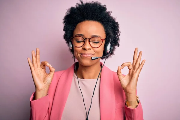 Young African American call center operator woman with curly hair using headset relax and smiling with eyes closed doing meditation gesture with fingers. Yoga concept.