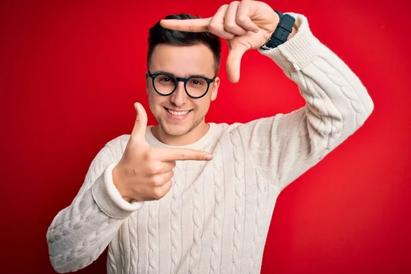 年轻英俊的高加索男子 戴着眼镜 穿着宽松的冬季毛衣 披着红色背景 微笑着 用双手和手指做框架 脸上洋溢着幸福的表情 创意和摄影概念 — 图库照片
