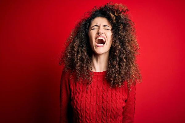 Young beautiful woman with curly hair and piercing wearing casual red sweater angry and mad screaming frustrated and furious, shouting with anger. Rage and aggressive concept.