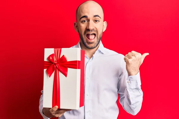 Jovem Careca Bonito Segurando Presente Apontando Polegar Até Lado Sorrindo — Fotografia de Stock