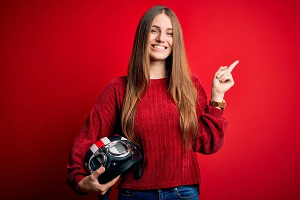 若いです美しいです赤毛Motocystist女性保持本ヘルメット以上赤背景非常に幸せなポインティングとともに手と指へ側 — ストック写真