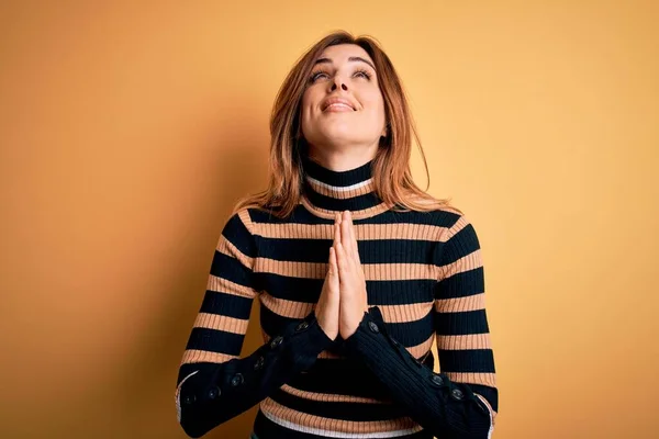 Young beautiful brunette woman wearing striped turtleneck sweater over yellow background begging and praying with hands together with hope expression on face very emotional and worried. Begging.