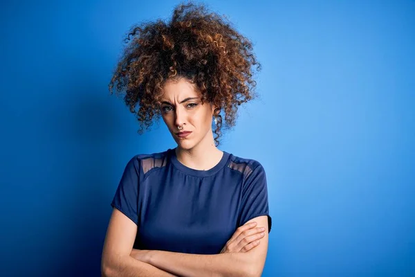 Young beautiful woman with curly hair and piercing wearing casual blue t-shirt skeptic and nervous, disapproving expression on face with crossed arms. Negative person.