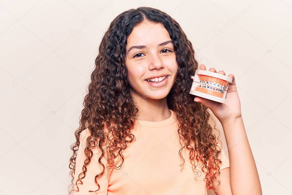 Beautiful kid girl with curly hair holding orthodontic looking positive and happy standing and smiling with a confident smile showing teeth 