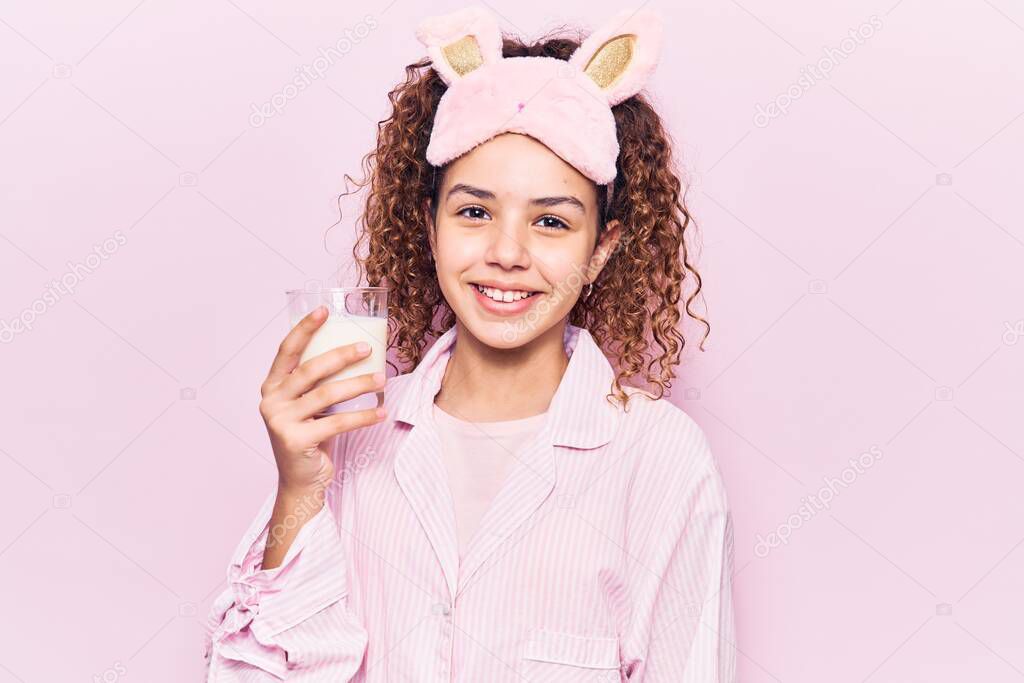 Beautiful kid girl with curly hair wearing sleep mask and pajama holding milk looking positive and happy standing and smiling with a confident smile showing teeth 