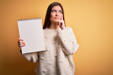 Mavi gözlü genç bir kadın sarı arka planda boş beyaz bir not defteri tutuyor. Ciddi bir yüz. Soru sormayı düşünüyor, kafası karışık bir fikir.