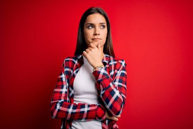 Genç, güzel, esmer, günlük gömlekler giyen bir kadın. İzole edilmiş kırmızı arka planda duruyor. Elleri çenesinde, soru sormayı düşünüyor. Düşünceli bir yüzle gülümsüyor. Şüphe kavramı.