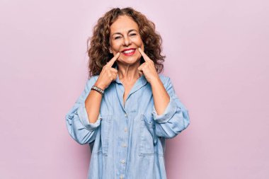 Orta yaşlı güzel bir kadın pembe arka planda sıradan bir kot gömlek giyiyor ağzı açık gülümsüyor, parmakları işaret ediyor ve zorla neşeli bir gülümseme sergiliyor.