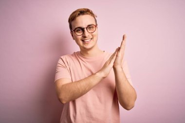 Genç, yakışıklı, günlük tişört giyen, pembe arka planda duran mutlu ve neşeli elleri alkışlayan, mutlu ve gururlu elleri bir arada tutan bir adam.