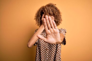 Genç, güzel, Afro-Amerikalı bir kadın, günlük gömleğini giymiş sarı arka plan üzerinde duruyor, gözleri ellerle örtüyor ve üzgün ve korku ifadeleriyle jestleri durduruyor. Utanç verici ve olumsuz bir kavram..