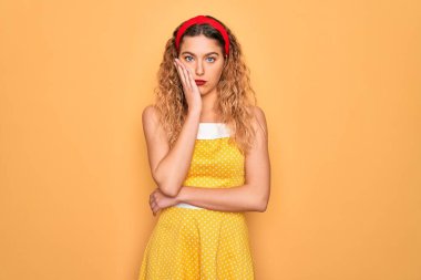 Sarı arka planda dikilip duran mavi gözlü güzel sarışın kadın kollarını kavuşturup depresyondan sıkılmış ve yorgun görünüyor..