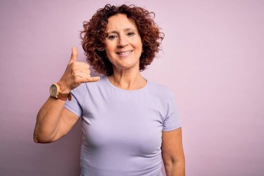 Orta yaşlı güzel kıvırcık saçlı kadın pembe arka planda rahat bir tişört giyip telefonla el ve parmaklarıyla, telefonla konuşur gibi gülümsüyor. İletişim kavramları.