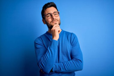 Mavi arka planda günlük süveter ve gözlük takan sakallı genç yakışıklı adam çapraz kollarla gülümseyen ve elleri çenesinde yükselen kameraya bakıyor. Olumlu düşünüyorum..