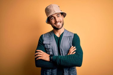 Tatilde sakallı yakışıklı turist sarı arka planda kaşif şapkası takıyor ve kollarını kavuşturup kameraya bakıyor. Pozitif kişi.