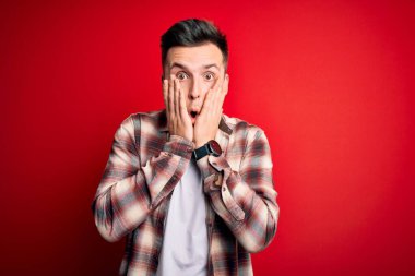 Kırmızı izole arka plan üzerine gündelik modern gömlek giyen genç beyaz adam korkmuş ve şok olmuş, şaşırmış ve şaşırmış bir yüz ifadesi var.