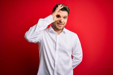 Mavi gözlü, zarif gömlekli genç bir iş adamı kırmızı izole edilmiş arka planda duruyor. El hareketleriyle gülümsüyor, gözleriyle mutlu yüzle parmaklarının arasından bakıyor..