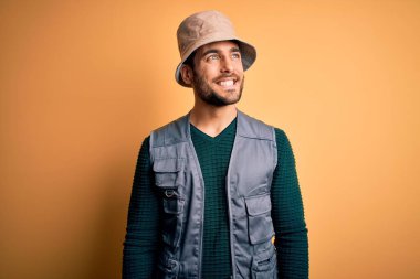 Tatilde sakallı yakışıklı bir turist sarı arka planda kaşif şapkası takıyor ve yüzünde gülümseme, doğal bir ifade ile diğer tarafa bakıyor. Kendinden emin gülen.